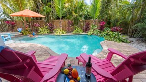 A backyard with a swimming pool, surrounded by tropical plants and a wooden fence. There are pink chairs and a table with fruit and a bottle.
