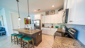 A modern kitchen with white cabinets, a granite countertop, and a built-in oven. There are two bar stools with green seats and a stainless steel refrigerator.