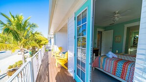 A balcony with wooden flooring, yellow chairs, and a bed with a patterned bedspread.
