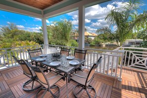 A covered outdoor dining area with a table set for four, surrounded by chairs, on a wooden deck with a view of greenery and a building in the background. - Canal-Front Oasis with Private Dock, Pool, Near Beach and Shops - Aqua Cove (Anna Maria)
