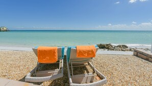 Two wicker lounge chairs with orange towels on a pebble beach, overlooking a calm sea and a clear sky. - Bayfront Home with Amazing Water Views! Private pool! – Breakaway (Anna Maria)