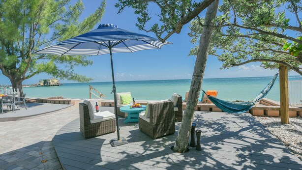 A wooden deck with wicker furniture, a hammock, and a blue-striped umbrella overlooking a beach. - Bayfront Home with Amazing Water Views! Private pool! – Breakaway (Anna Maria)