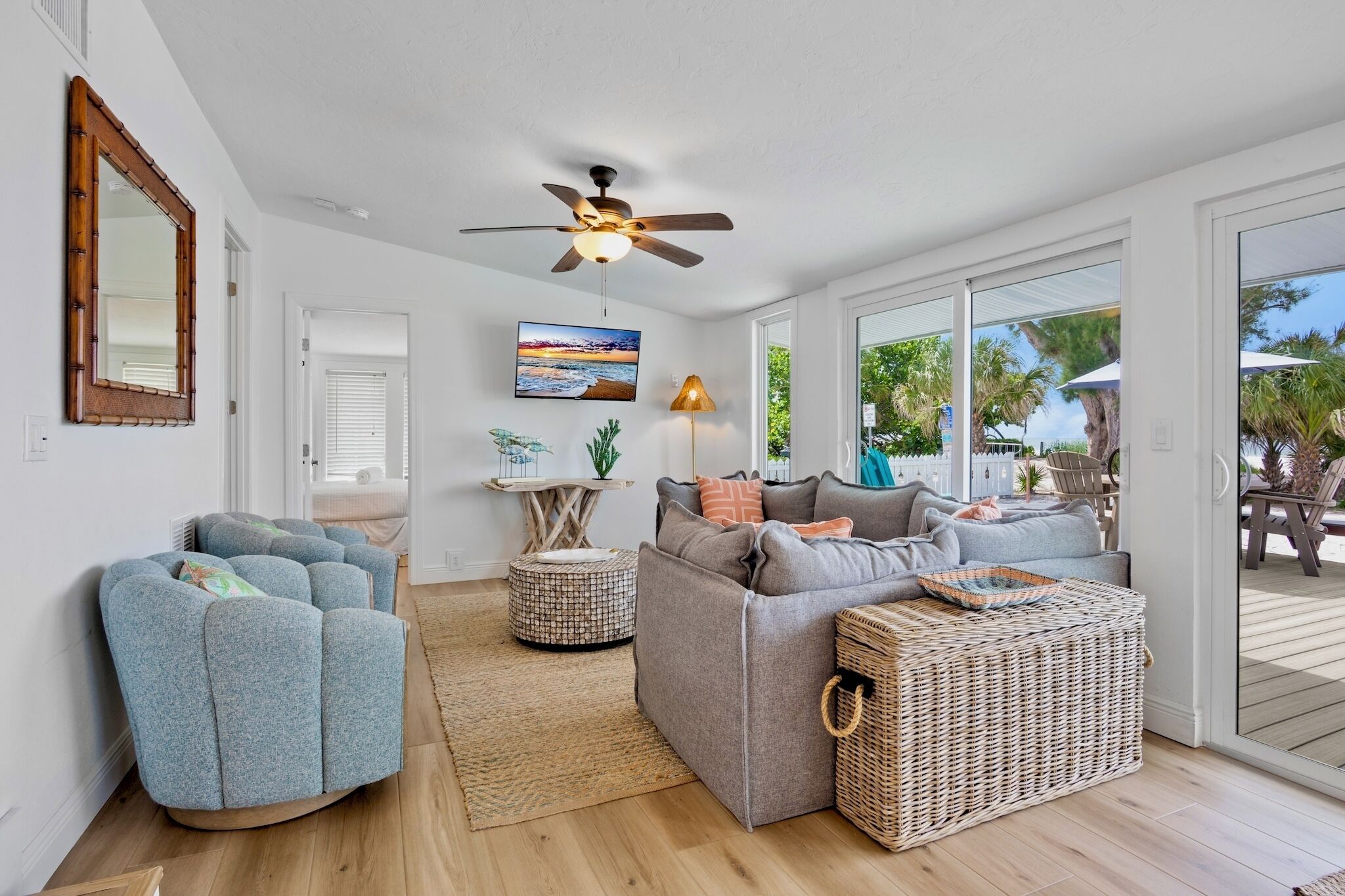 A spacious living room with a gray sofa, blue armchair, and wicker ottoman. A ceiling fan and a TV are mounted on the wall.