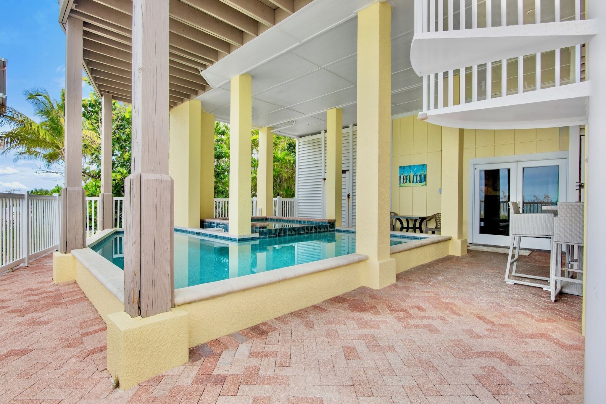 A covered outdoor pool area with a pool, white railings, and a patio with a table and chairs.
