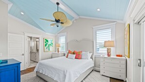 Une chambre à coucher comprenant un lit, des tables de chevet, une commode, un ventilateur de plafond et une fenêtre avec des stores.