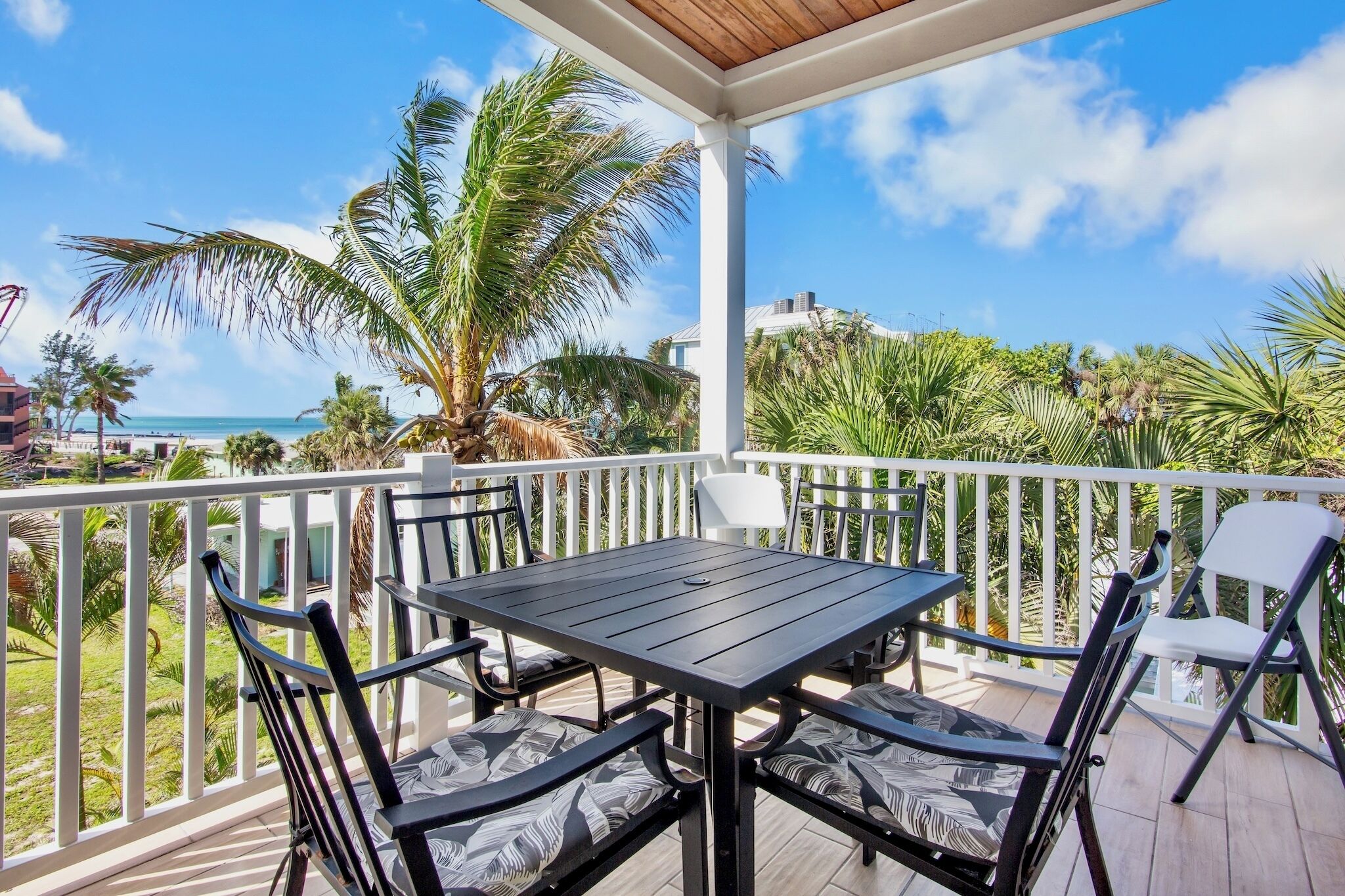 A covered outdoor area with a table and chairs, a white railing, and a view of the beach and palm trees.