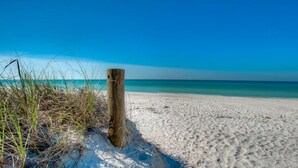 Une plage de sable avec un poteau en bois et de l’herbe au premier plan.