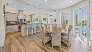 A spacious kitchen with wooden flooring, a dining area with wicker chairs, and large windows. - Sunset Views from Rooftop, Heated Pool, Game Room- Caribbean Dream (Holmes Beach)