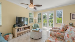A living room with a television, a sofa, a chair, a round ottoman, and large windows with a view of greenery.