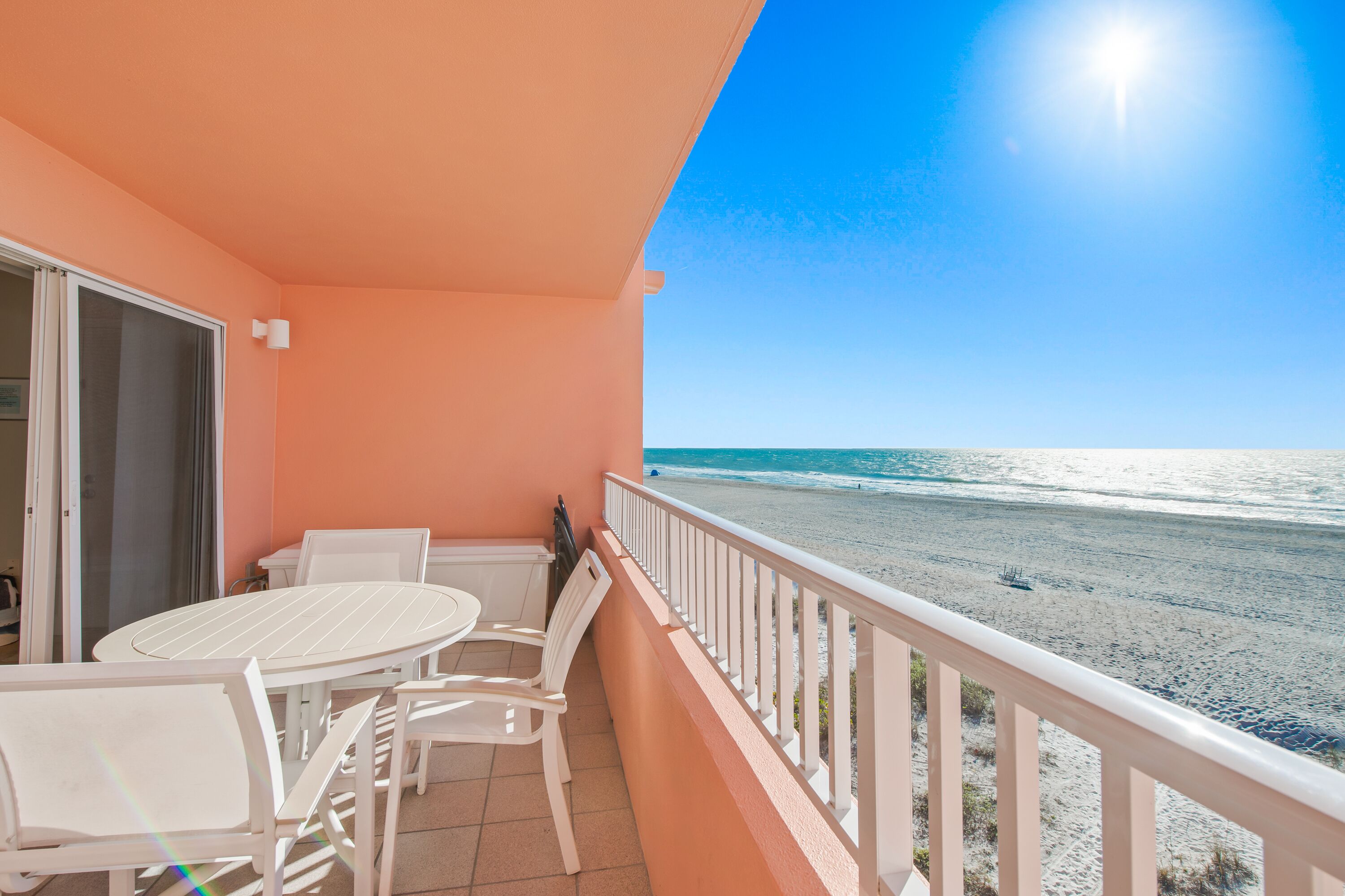A balcony with a white table and chairs, overlooking a beach with the ocean and a clear sky.