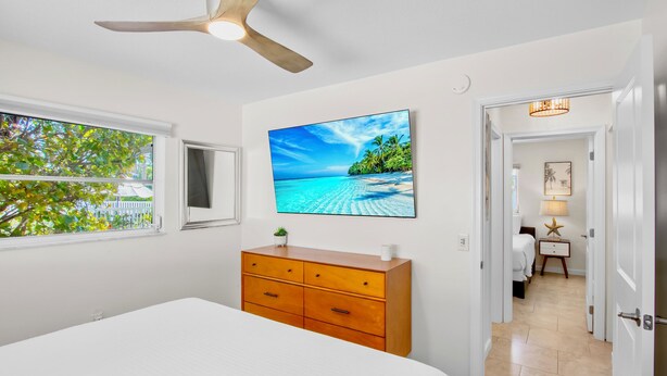 A bedroom with a bed, a wooden dresser, a ceiling fan, a TV on the wall, and a window with a view of trees. - Cozy Family Duplex with Pool! Close to the Gulf Beaches (Anna Maria)