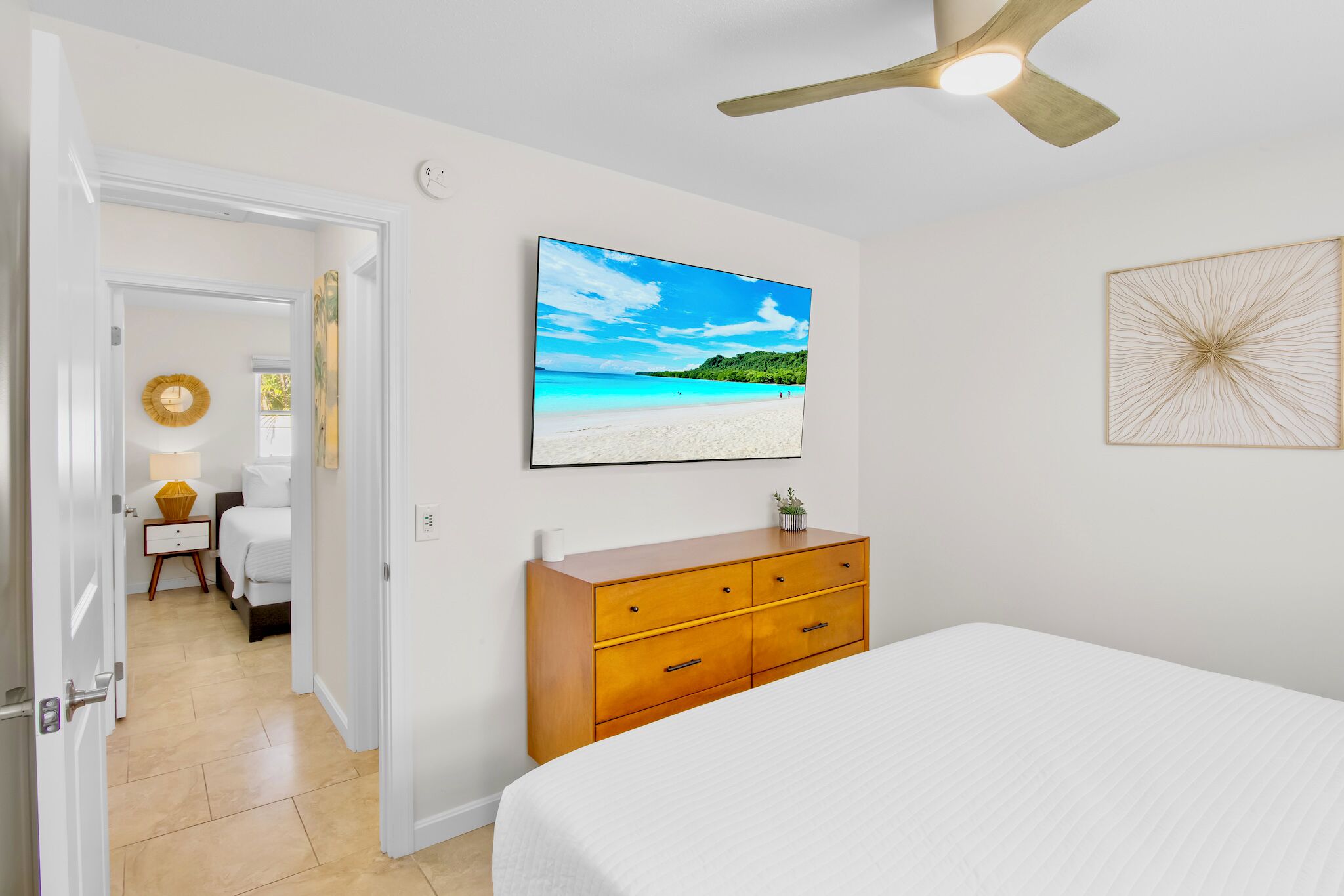 A bedroom with a bed, a wooden dresser, a ceiling fan, a wall-mounted TV displaying a beach scene, and a door leading to another room.