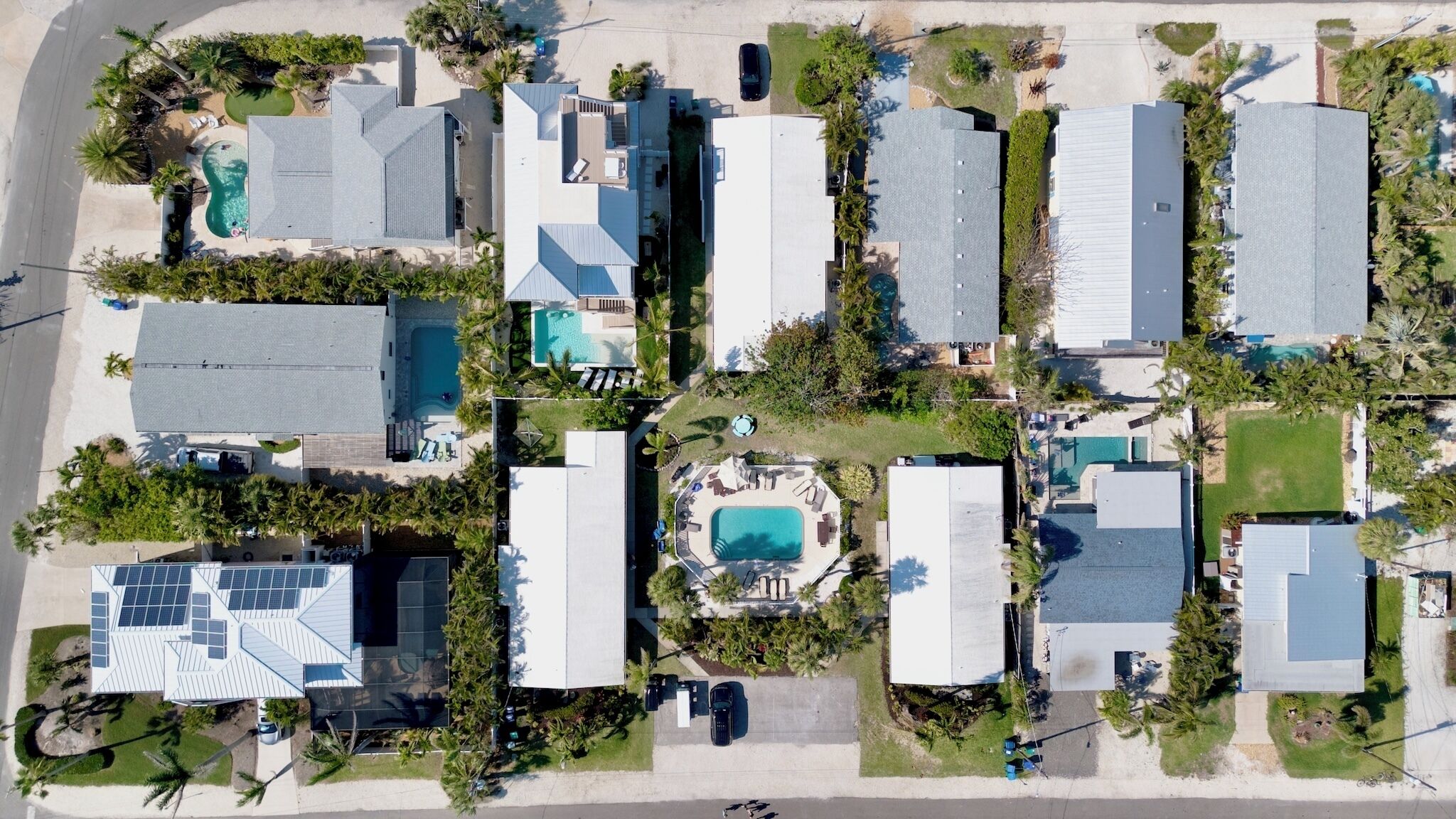 An aerial view of a residential area with multiple houses, each with a swimming pool and surrounded by palm trees.