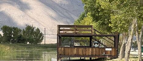 Une terrasse en bois au design rustique, donnant sur un lac paisible, entourée d’une végétation luxuriante et bordée de montagnes.