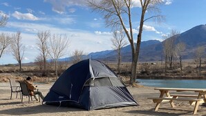 Un campeggio con una tenda, sedie e un tavolo da picnic lungo un fiume, sullo sfondo montagne.