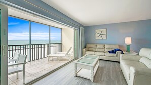 A spacious balcony with a view of the ocean, a white sofa, a glass-top coffee table, and a lamp on a side table. - Beachfront Condo with Amazing Views and Pool! Near Bridge Street! (Bradenton Beach)