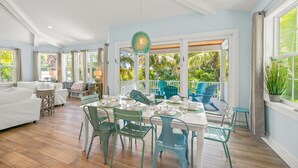 A spacious room with a dining area, white furniture, and large windows offering a view of the outdoors.