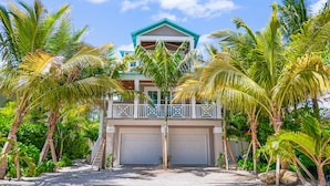 A two-story house with a balcony, surrounded by palm trees and a gravel driveway.