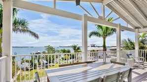 A covered outdoor area with a wooden table and chairs, white railings, and a view of the sea and distant buildings. - Bayblue — Spacious 6BR Retreat with Pool and Dock! (Bradenton Beach)
