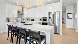A modern kitchen with white cabinetry, a large island with bar stools, stainless steel appliances, and woven pendant lights. - Bayblue — Spacious 6BR Retreat with Pool and Dock! (Bradenton Beach)