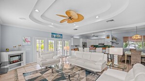 A spacious living room with a white sofa, a glass coffee table, and a fireplace. There is a kitchen area with white cabinets and a dining table with chairs. - Private Home with Heated Pool, Hot Tub, and Balconies! Near Beach and Dining (Anna Maria)