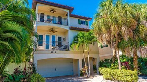 A two-story building with a balcony on the upper floor, a garage, and surrounded by palm trees and other vegetation.