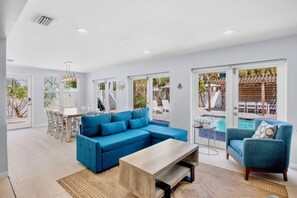 A spacious living area with a blue sectional sofa, a wooden coffee table, and a dining table with chairs. There is a glass door leading to a pool area with lounge chairs.