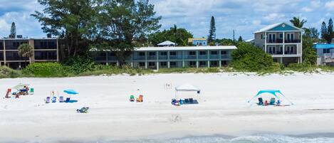 Ein Strand mit mehreren Liegestühlen und Sonnenschirmen, einem Zelt und einem Gebäude mit mehreren Balkonen.