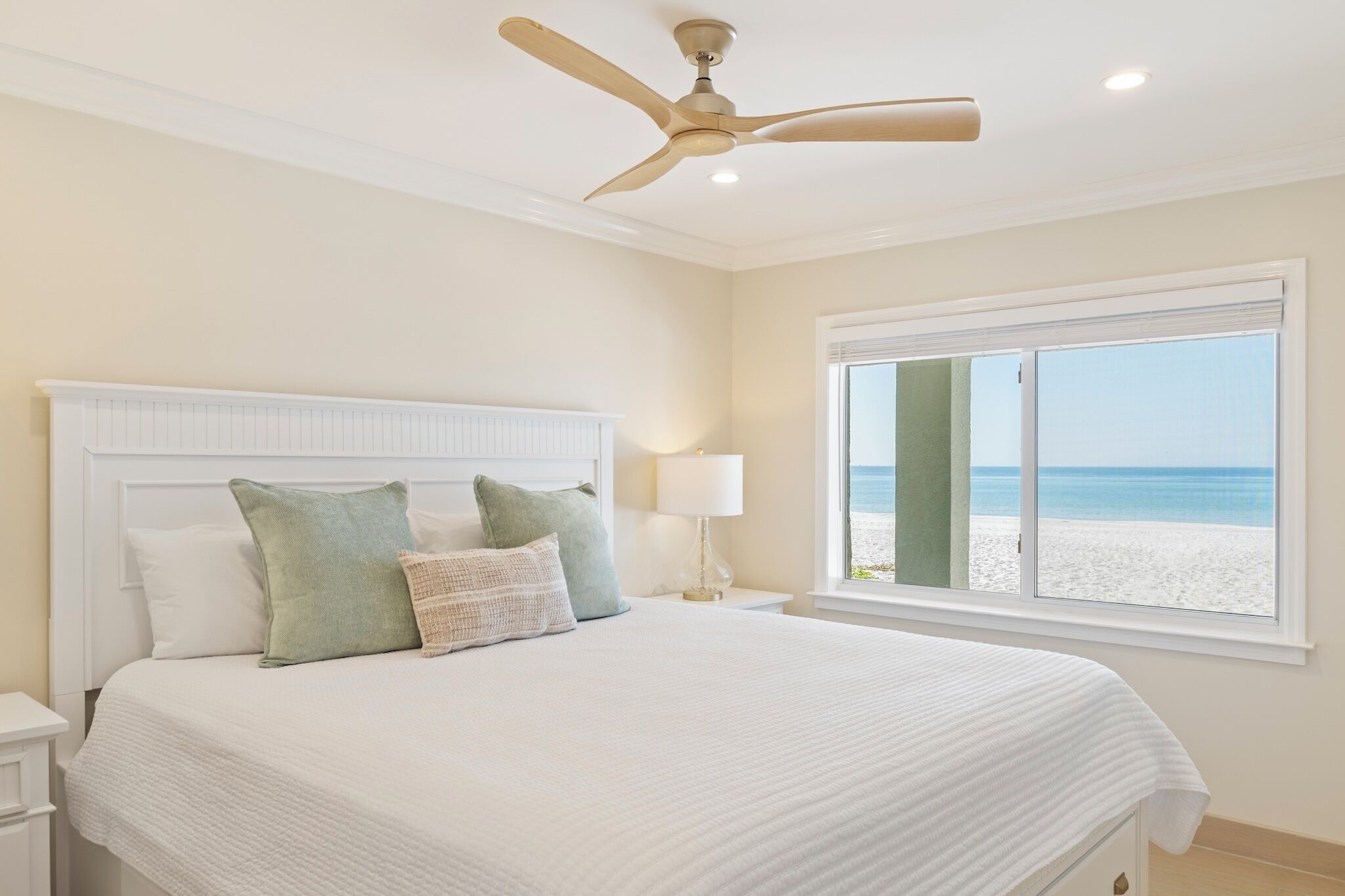 A bedroom with a large bed, a ceiling fan, a nightstand, a lamp, and a window with a beach view.