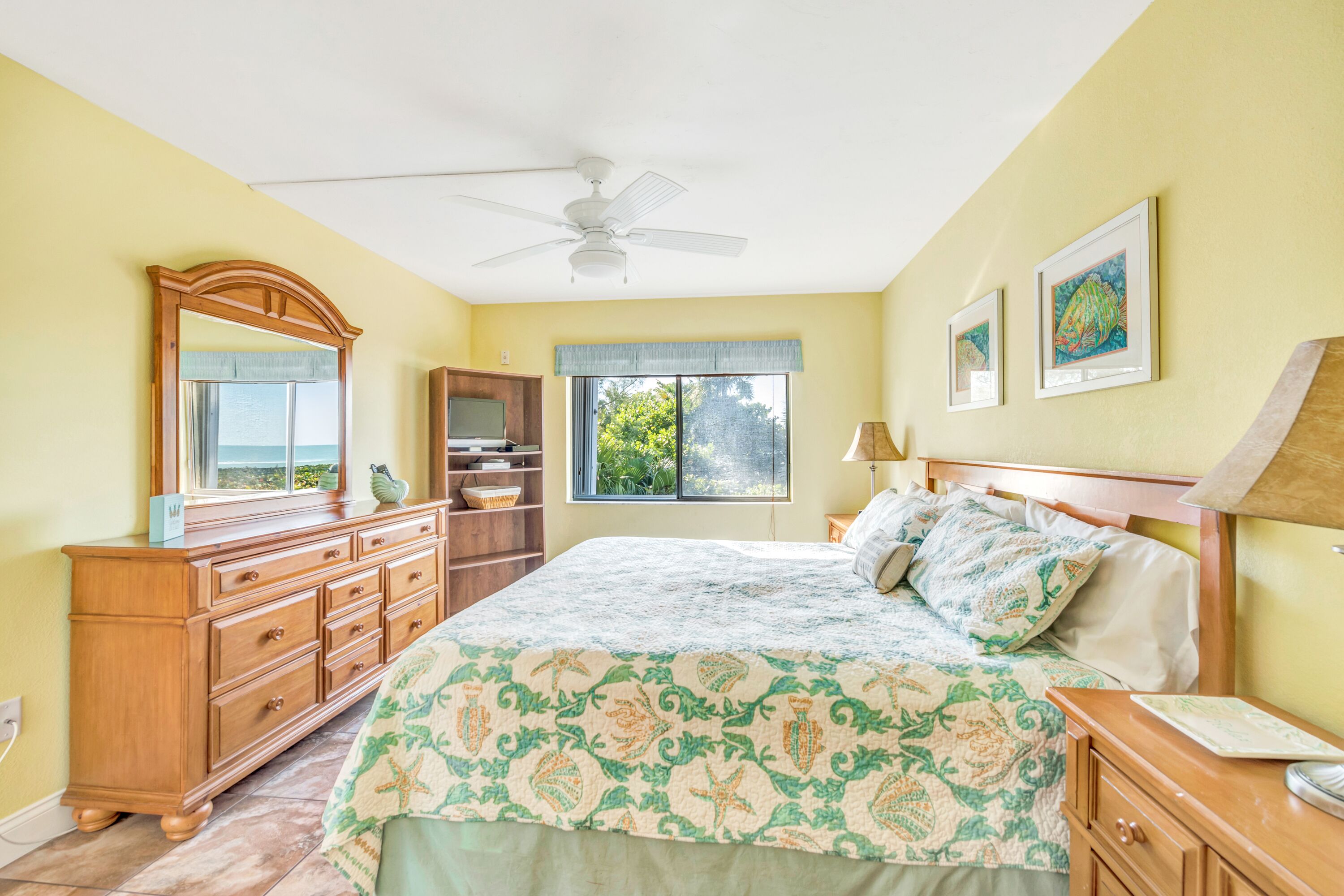 Un dormitorio con cama, cómoda, tocador, ventilador de techo, ventana con vistas y obras de arte en la pared.