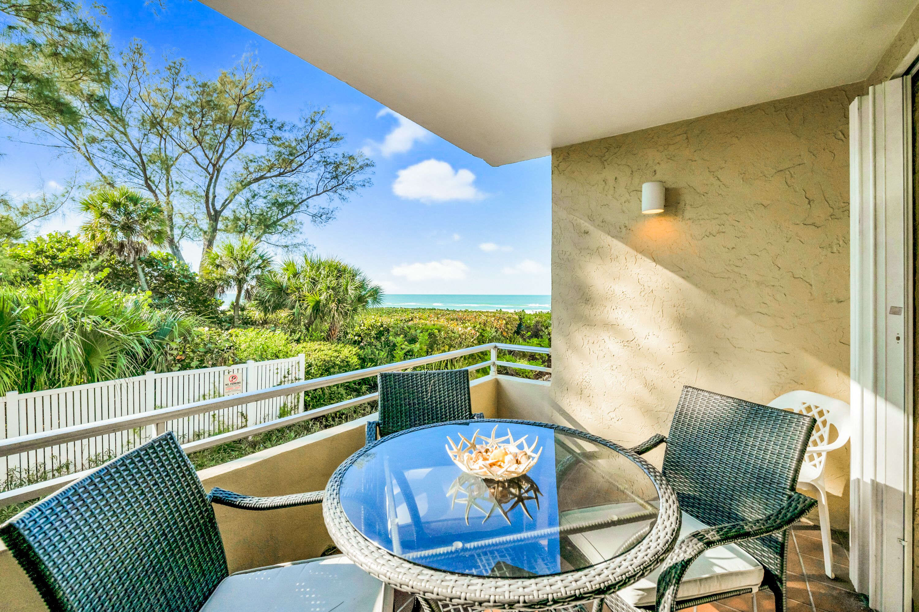 A balcony with a glass table and chairs, overlooking a beach and lush greenery.
