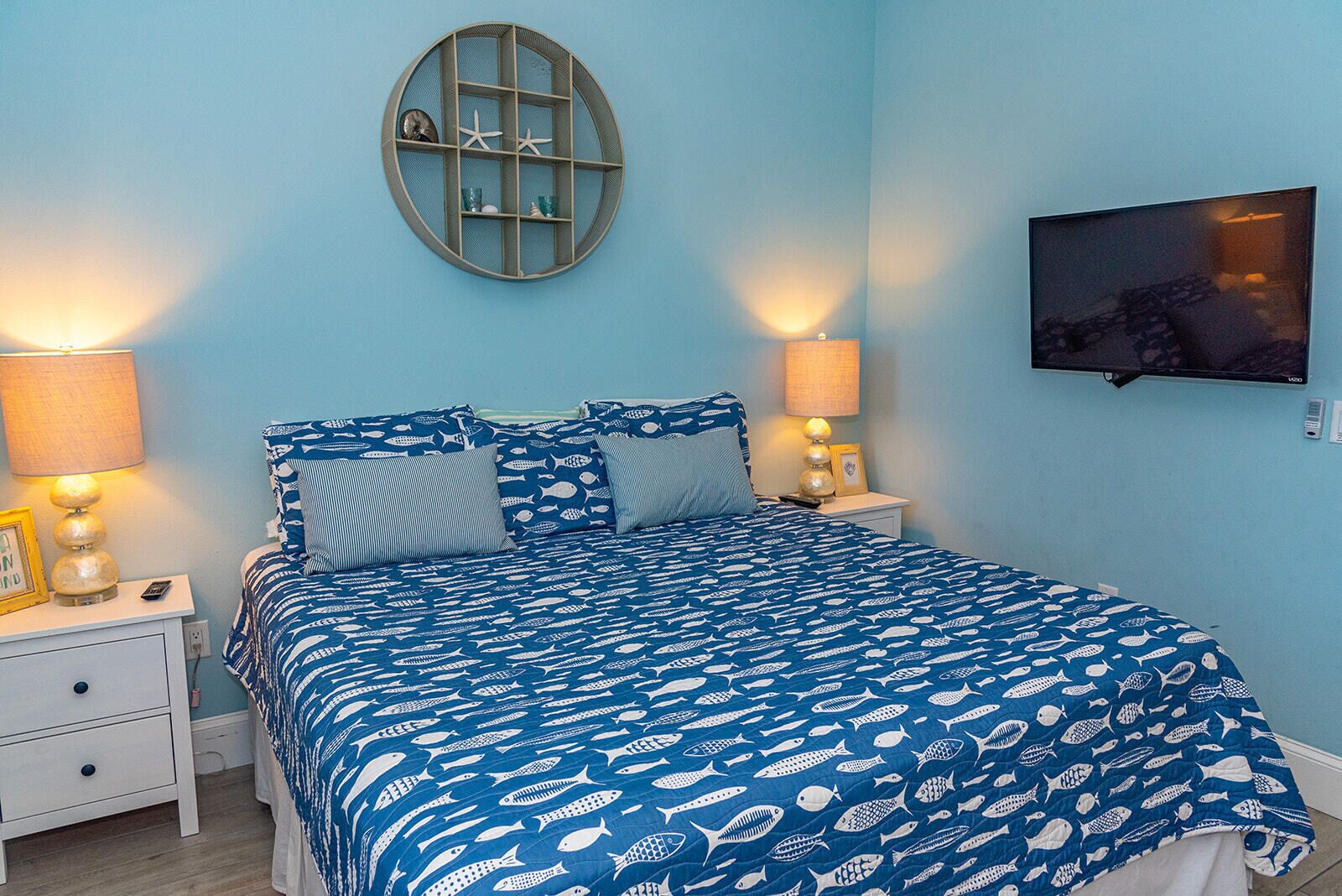 A bedroom with a blue bedspread, two bedside tables with lamps, a round wall shelf, and a TV mounted on the wall.