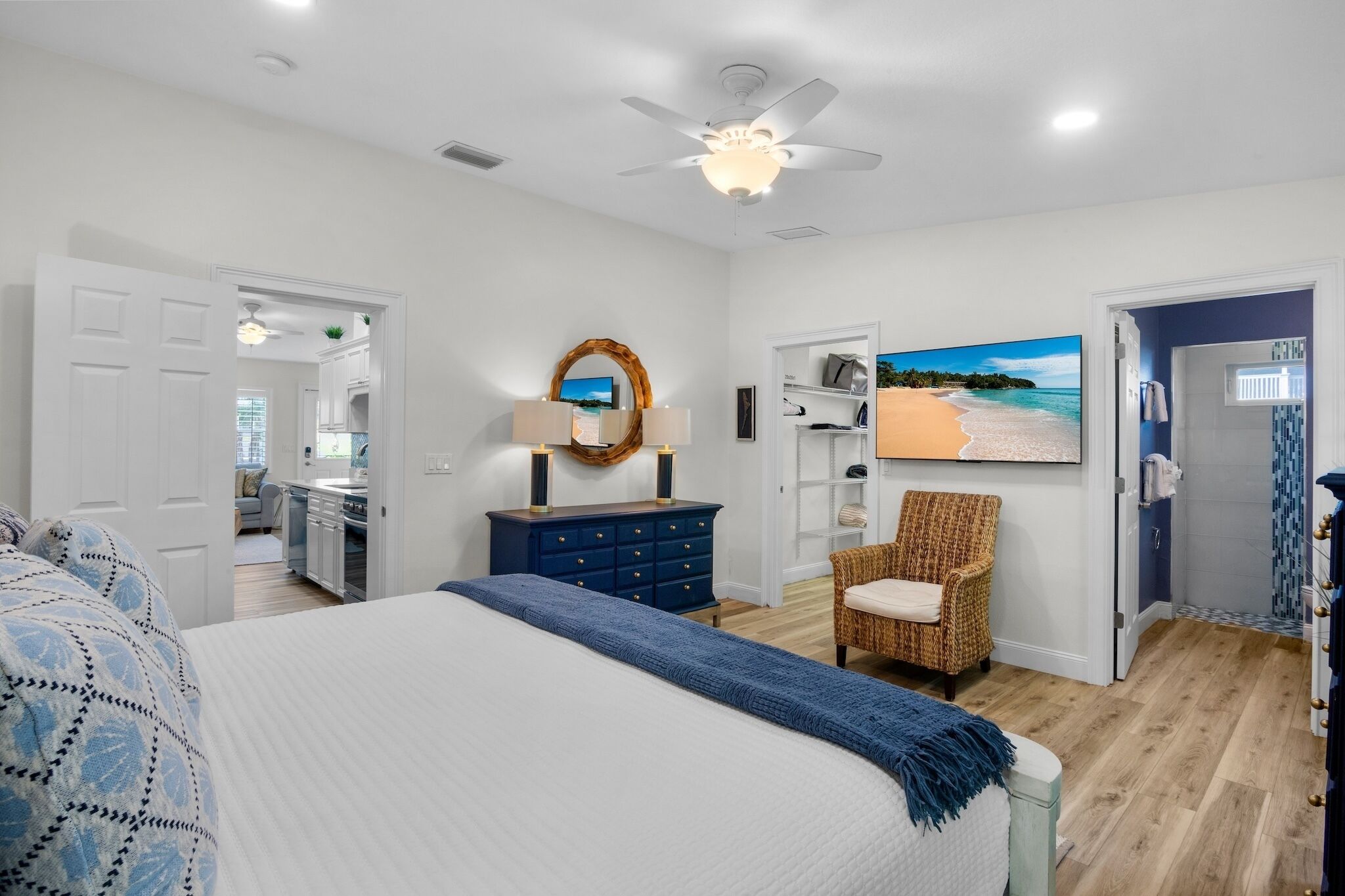 A bedroom with a bed, a dresser, a chair, a ceiling fan, and a painting of a beach.