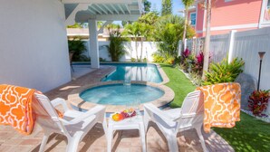 A backyard with a swimming pool, two white plastic chairs with orange covers, and a table with fruits and glasses.