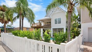 A two-story house with a balcony, surrounded by a white fence and palm trees. - Large Private Beach Home! Heated Pool, Elevator, Balcony, and Near the Beach! (Holmes Beach)