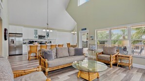 A spacious living area with a sofa, chairs, and a glass coffee table. A kitchen with white cabinets and appliances is visible in the background. - Large Private Beach Home! Heated Pool, Elevator, Balcony, and Near the Beach! (Holmes Beach)