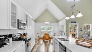 A modern kitchen with white cabinetry, a central island, and a dining area with wicker chairs. - Large Private Beach Home! Heated Pool, Elevator, Balcony, and Near the Beach! (Holmes Beach)