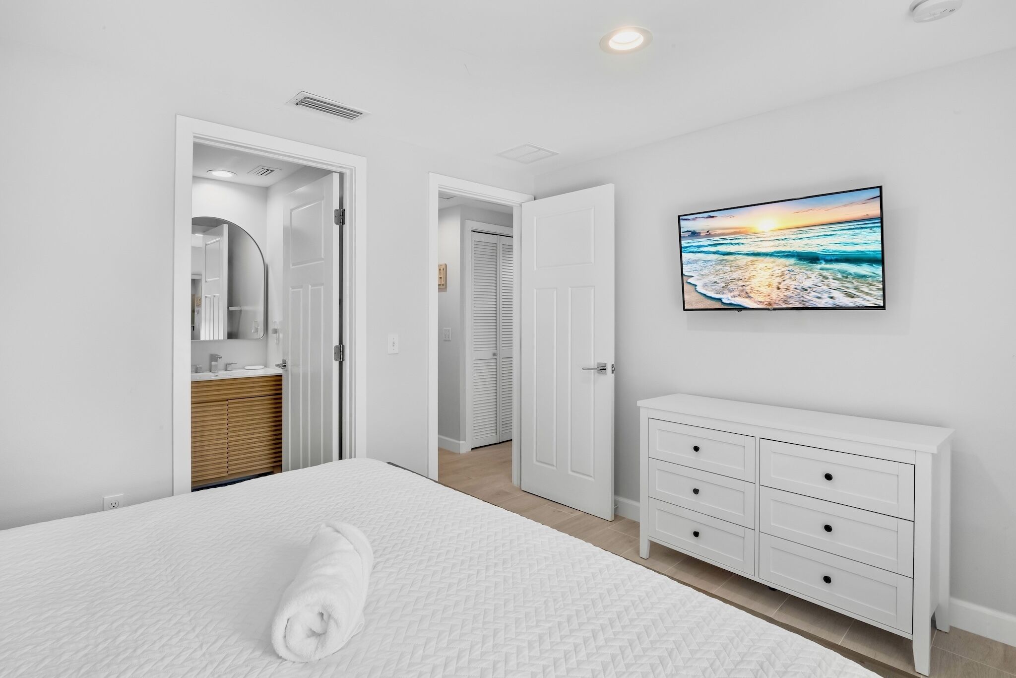 Un dormitorio con cama, cómoda blanca, un cuadro de una playa y un baño visible a través de una puerta abierta.
