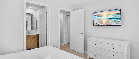 Un dormitorio con cama, cómoda blanca, un cuadro de una playa y un baño visible a través de una puerta abierta.