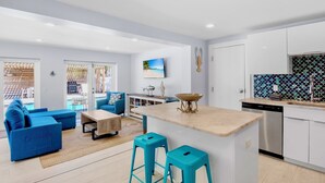 Un salon moderne avec un canapé bleu, une table basse et un coin cuisine équipé d’un lave-vaisselle et doté d’un plan de travail recouvert de carreaux.