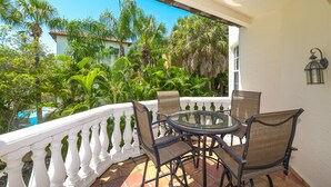 Un balcón con mesa y sillas de vidrio, rodeado por una barandilla blanca, con vistas a un exuberante jardín con palmeras y una piscina.