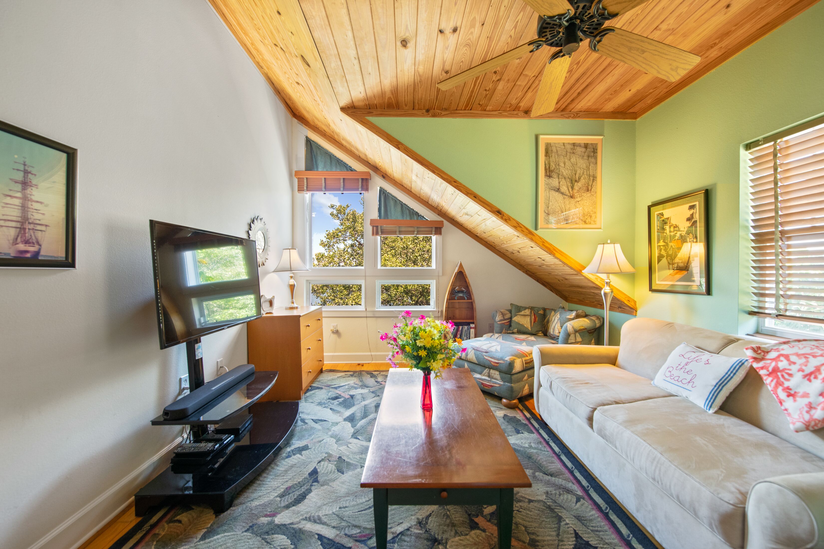 A cozy living room with a wooden ceiling, a flat-screen TV, a coffee table with flowers, a sofa, and a window with blinds.
