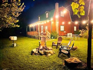 A cozy outdoor seating area with a fire pit, surrounded by chairs and a stone fire ring, in front of a house with a porch and string lights. - Private Cabin: Mountain Views & Pond!🎣 (Barnardsville)