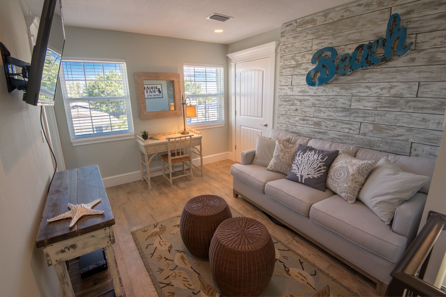 A living room with a beige sofa, a wooden coffee table, a desk with a chair, and a wall-mounted TV.