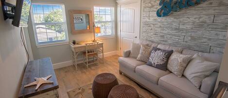 A living room with a beige sofa, a wooden coffee table, a desk with a chair, and a wall-mounted TV.