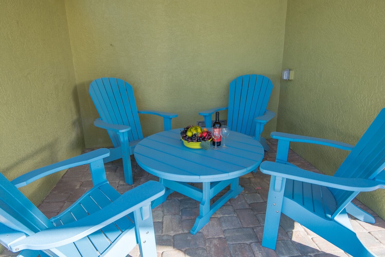 A blue table and four matching chairs arranged around it, with a bowl of fruit and a bottle of wine on the table.
