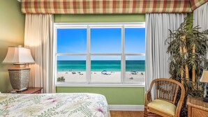 Ein Schlafzimmer mit großem Fenster und Blick auf den Strand, einem Bett mit Blumenmuster, einem Korbstuhl und einer Lampe auf einem Nachttisch.