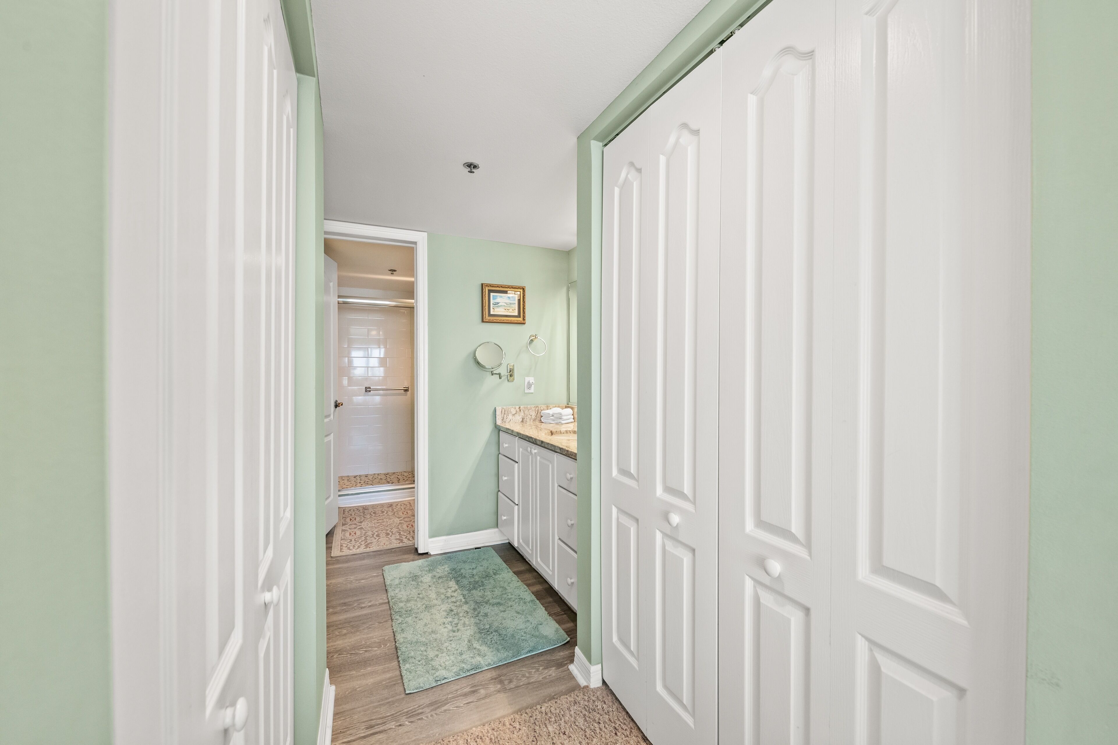 A narrow hallway with a light green wall, white doors, and a small bathroom with a sink and mirror.
