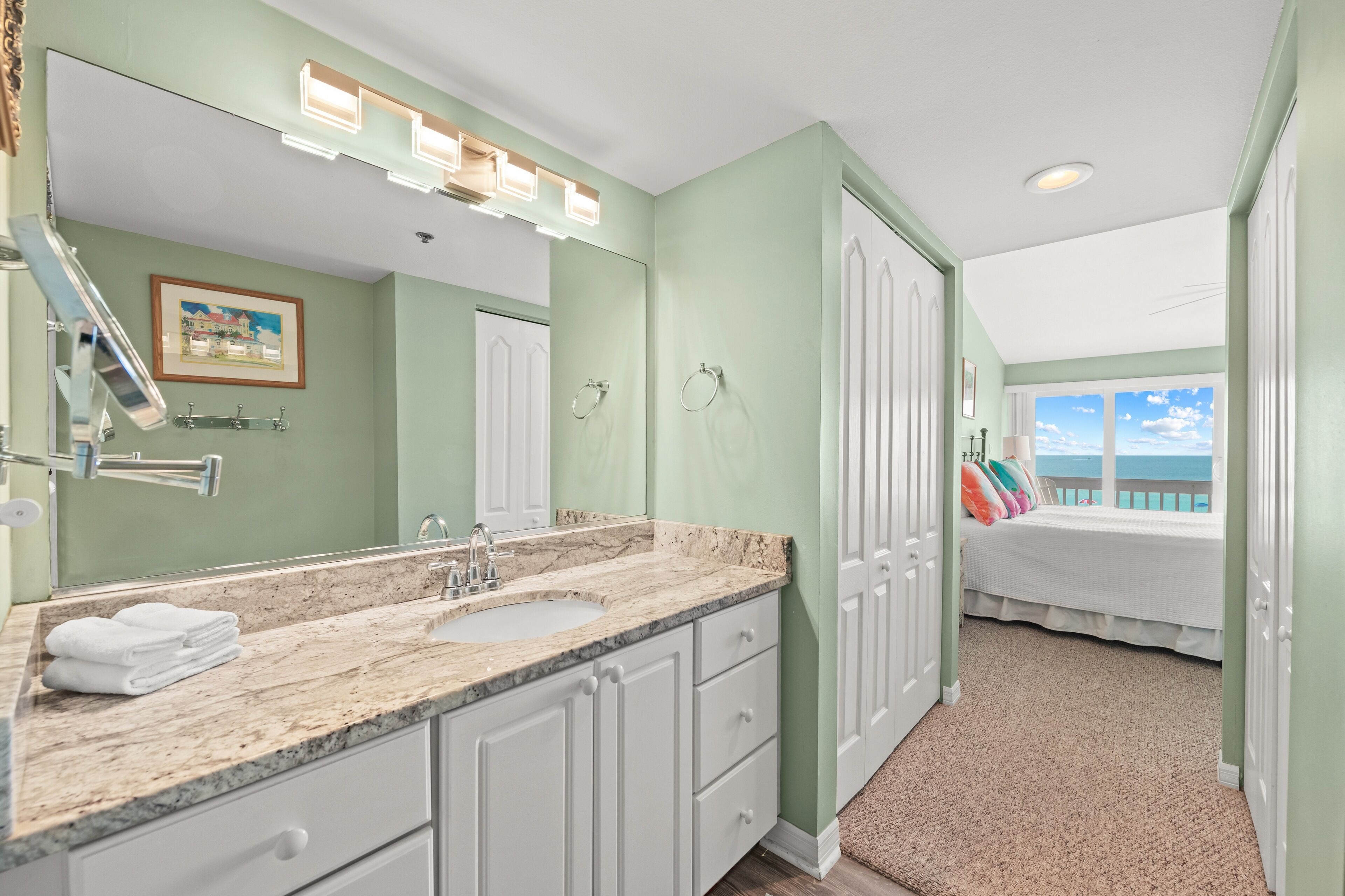 A bathroom with a double sink, white cabinets, and a large mirror. There is a framed picture on the wall and a window with a view of the ocean.