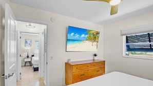 A bedroom with a bed, a wooden dresser, a painting of a beach, and a window with blinds.